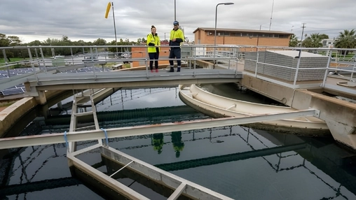 Two workers in hi-vis jackets on a platform above large circular water treatment tanks at an outdoor facility