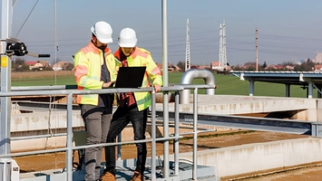 Two engineers wearing hard hats and hi-vis jackets review plans on a laptop at a water treatment facility