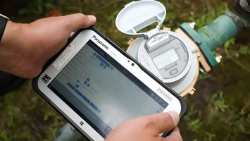 Technician using a rugged Panasonic tablet to read a smart water meter installed on a green pipe outdoors