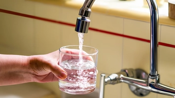 Person filling a clear glass with running tap water from a chrome kitchen faucet with tiled splashback