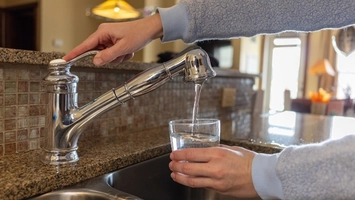Person filling a glass of water at a chrome kitchen tap over a dark granite sink in a domestic interior